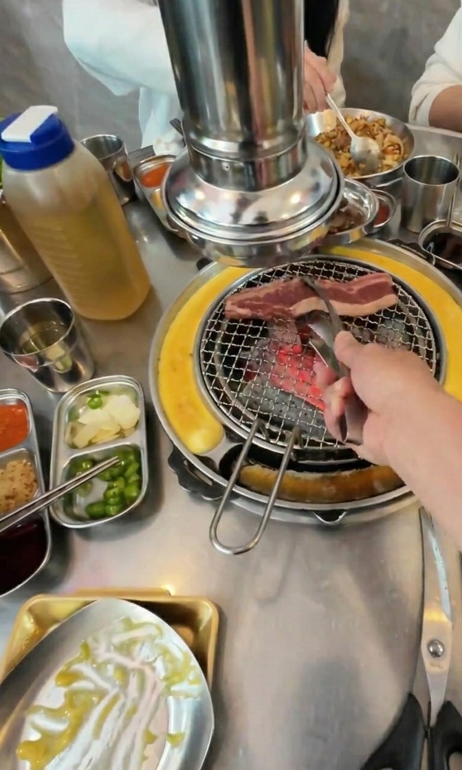 Une main déposant des lamelles de bœuf sur un gril à charbon entouré de petits plats daccompagnement.