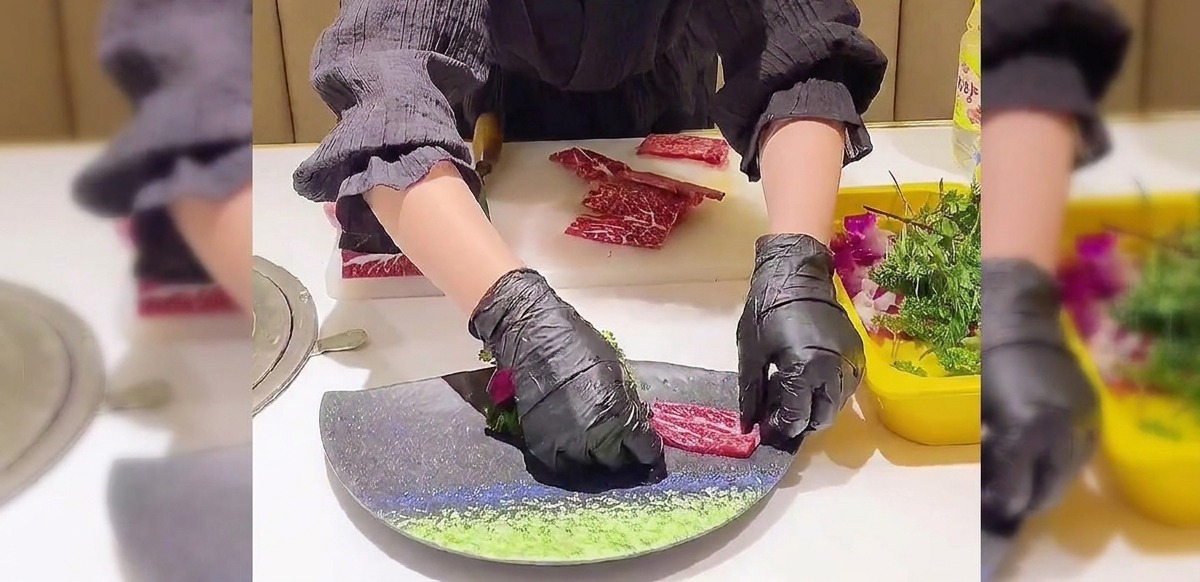 Mains gantées disposant délicatement des tranches de bœuf persillées en éventail sur une assiette sombre incurvée.