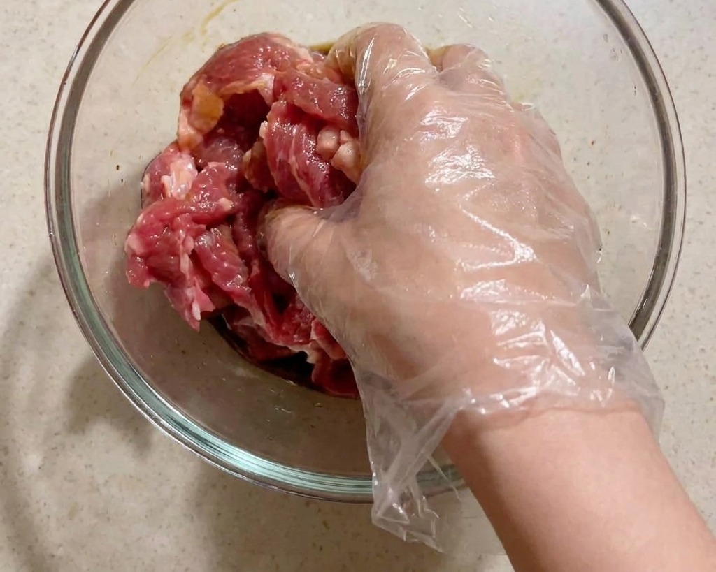 Une main gantée mélangeant vigoureusement les tranches de porc avec les assaisonnements liquides.
