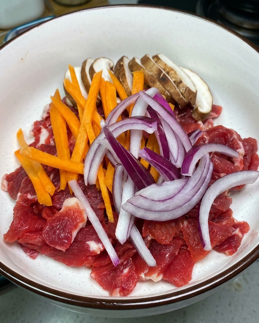 Un bol blanc rempli de tranches de bœuf rouge cru garni de lamelles de champignons shiitake, de carottes orange et doignons rouges.