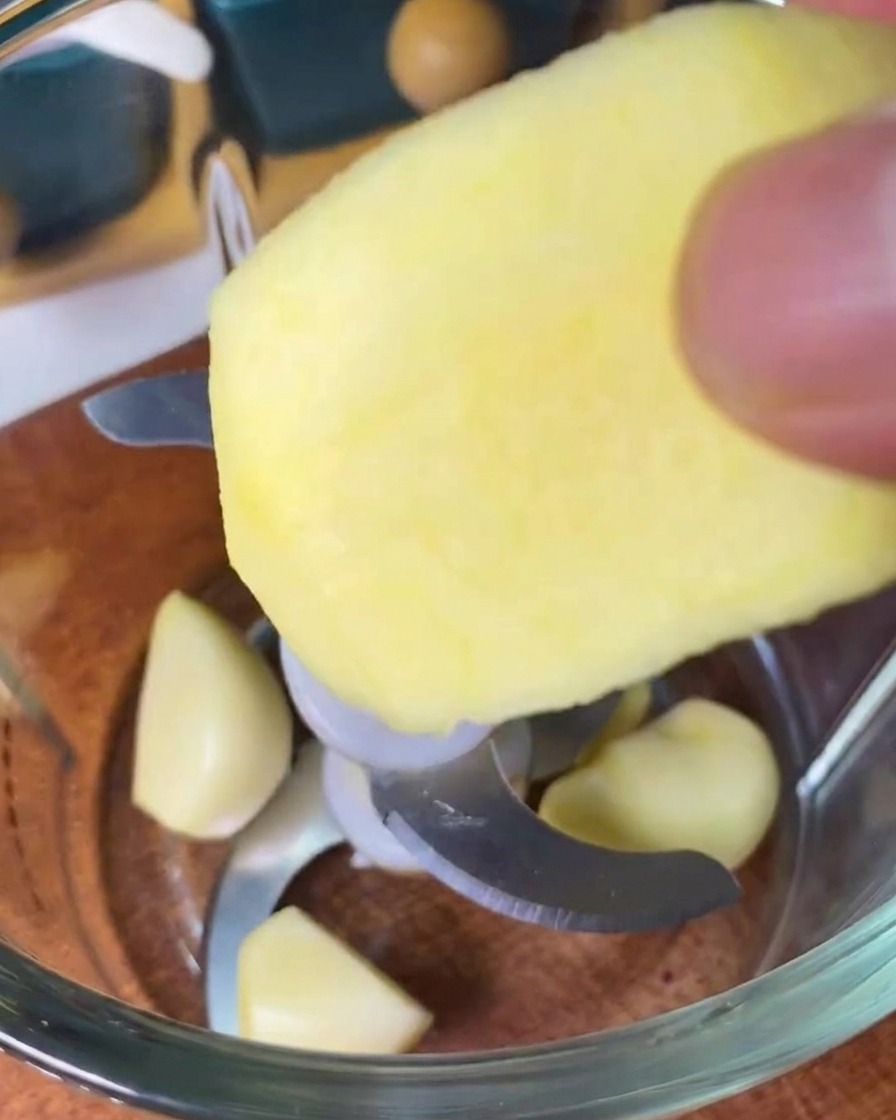 Une personne déposant une tranche de pomme pelée dans un mixeur contenant déjà plusieurs gousses dail entières.