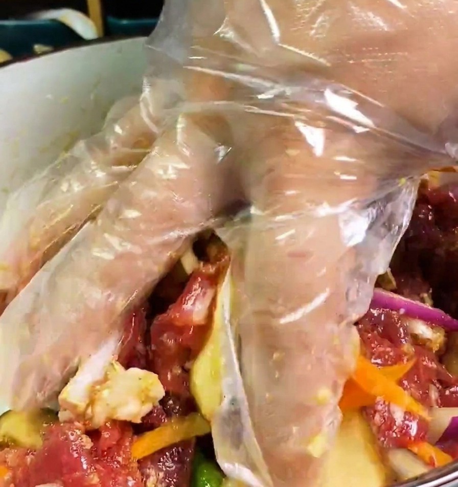 Une main gantée mélangeant du bœuf cru, des oignons, des carottes et des champignons dans un bol de marinade sombre.
