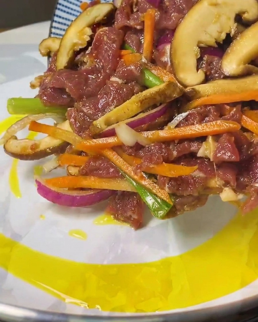 Lamelles de bœuf mariné et de légumes versées depuis une assiette à motifs bleus dans une poêle avec de lhuile chaude.