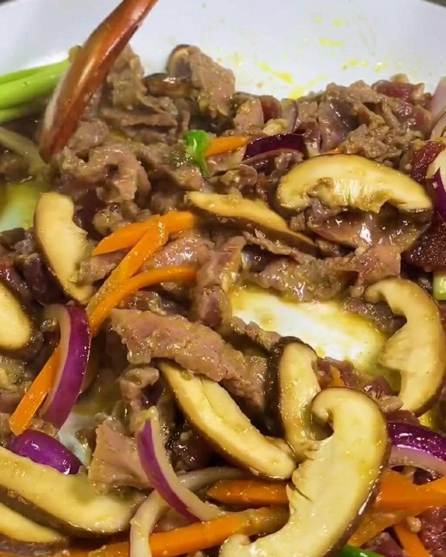 Fines tranches de bœuf, champignons shiitake et carottes sautés dans une poêle blanche avec une spatule en bois.