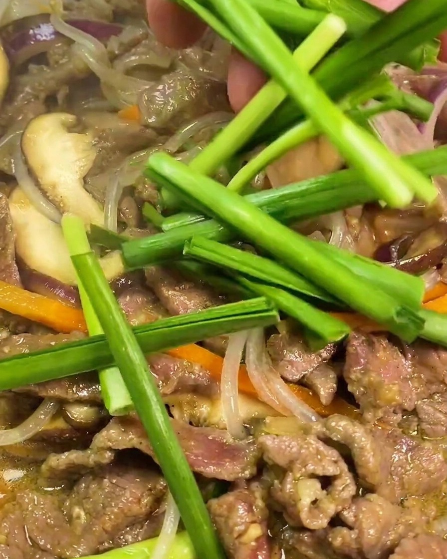 Segments de ciboule chinoise verte ajoutés à la main dans une poêle chaude contenant du bœuf sauté, des nouilles translucides, des carottes et des oignons.