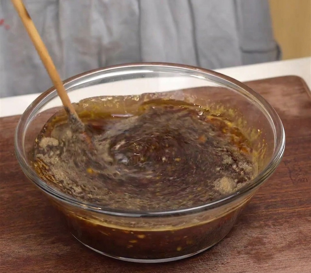 Une cuillère en bois mélange une marinade sombre et aromatique dans un bol en verre transparent.