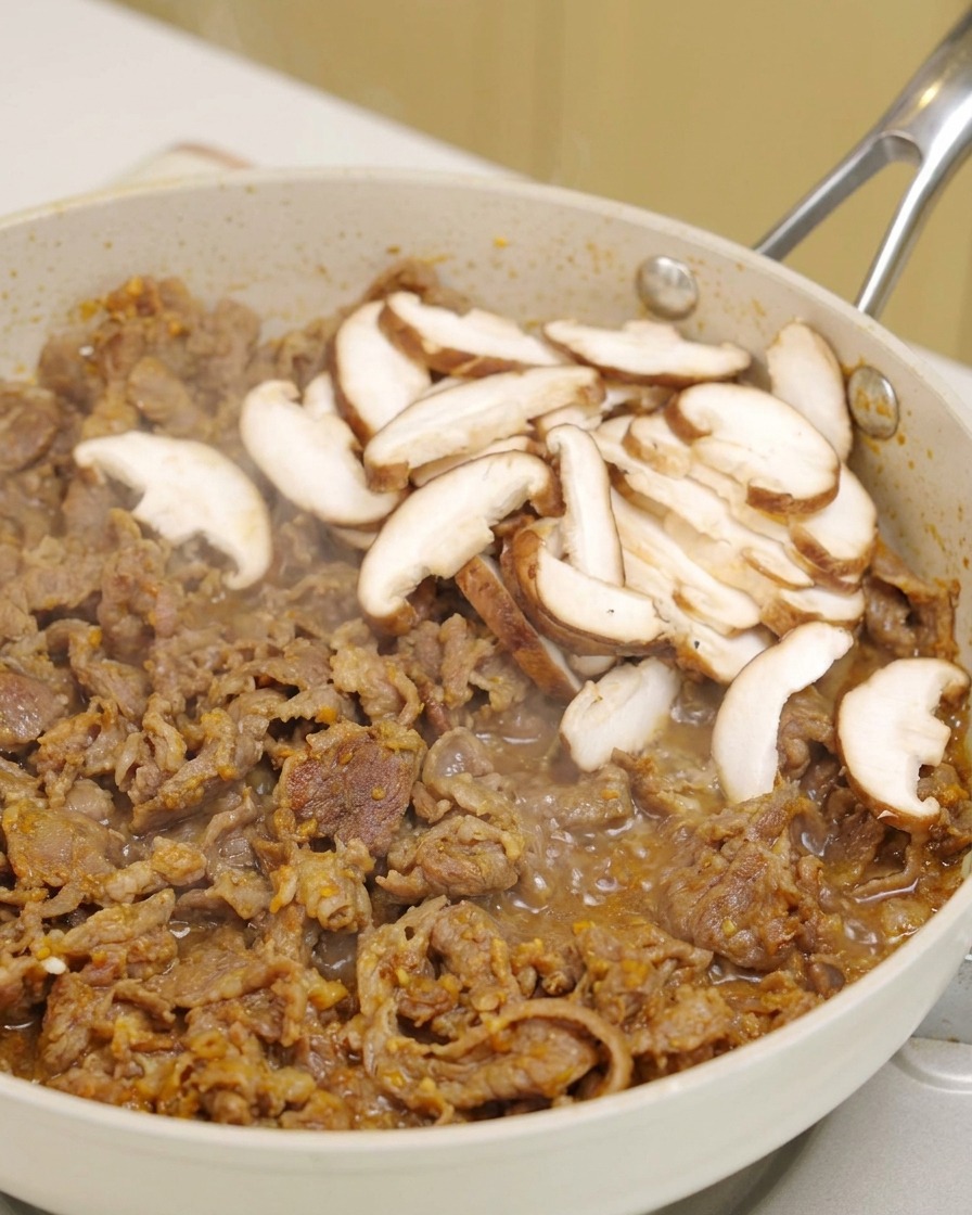 Des tranches de shiitakés frais déposées sur le bœuf grésillant dans une poêle blanche.