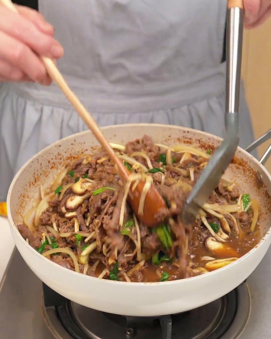 Une poêle remplie de bœuf cuit, champignons, oignons et cébettes mélangés avec une cuillère et une pince.