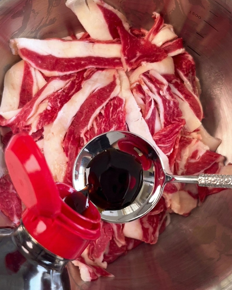 Sauce soja foncée versée à laide dune cuillère en métal sur un tas de tranches de bœuf cru dans un bol argenté.