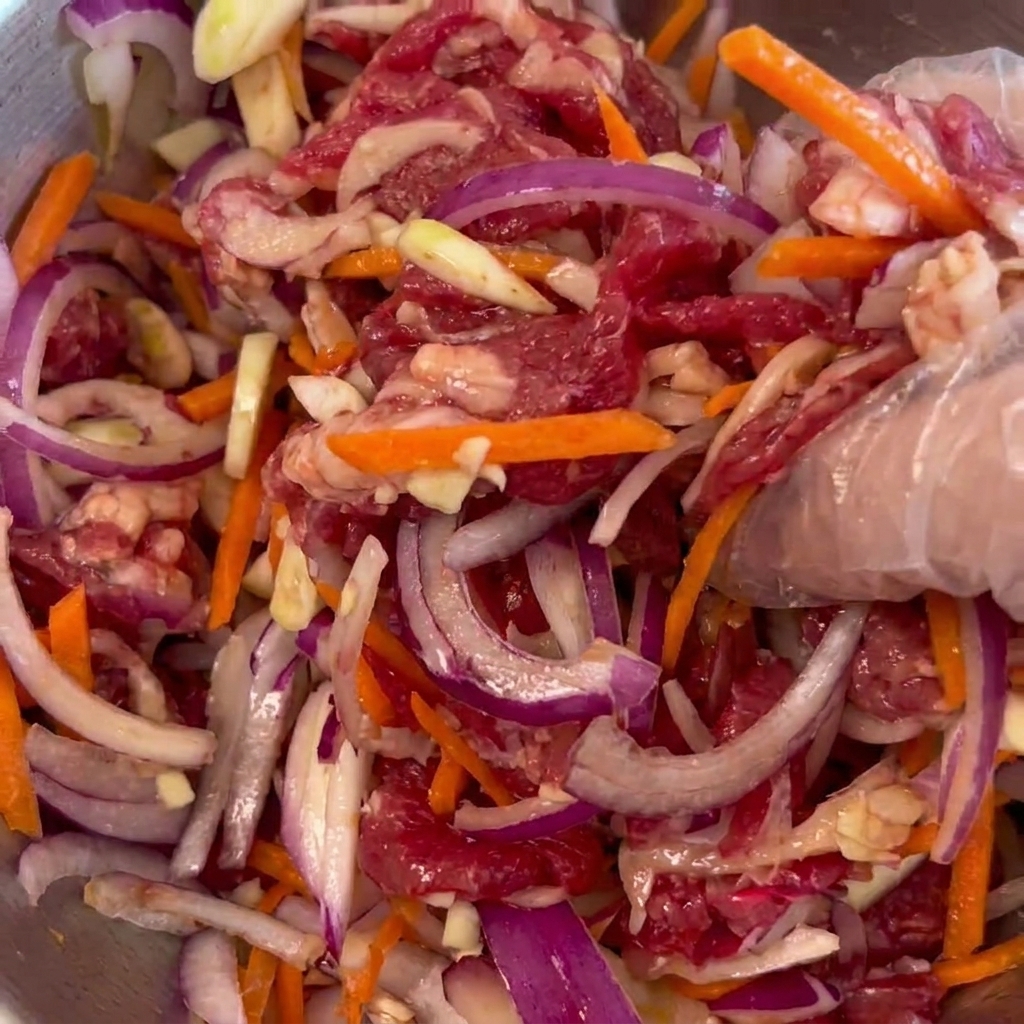 Une main portant un gant en plastique mélangeant du bœuf cru, des oignons rouges et des bâtonnets de carottes dans un bol en métal.
