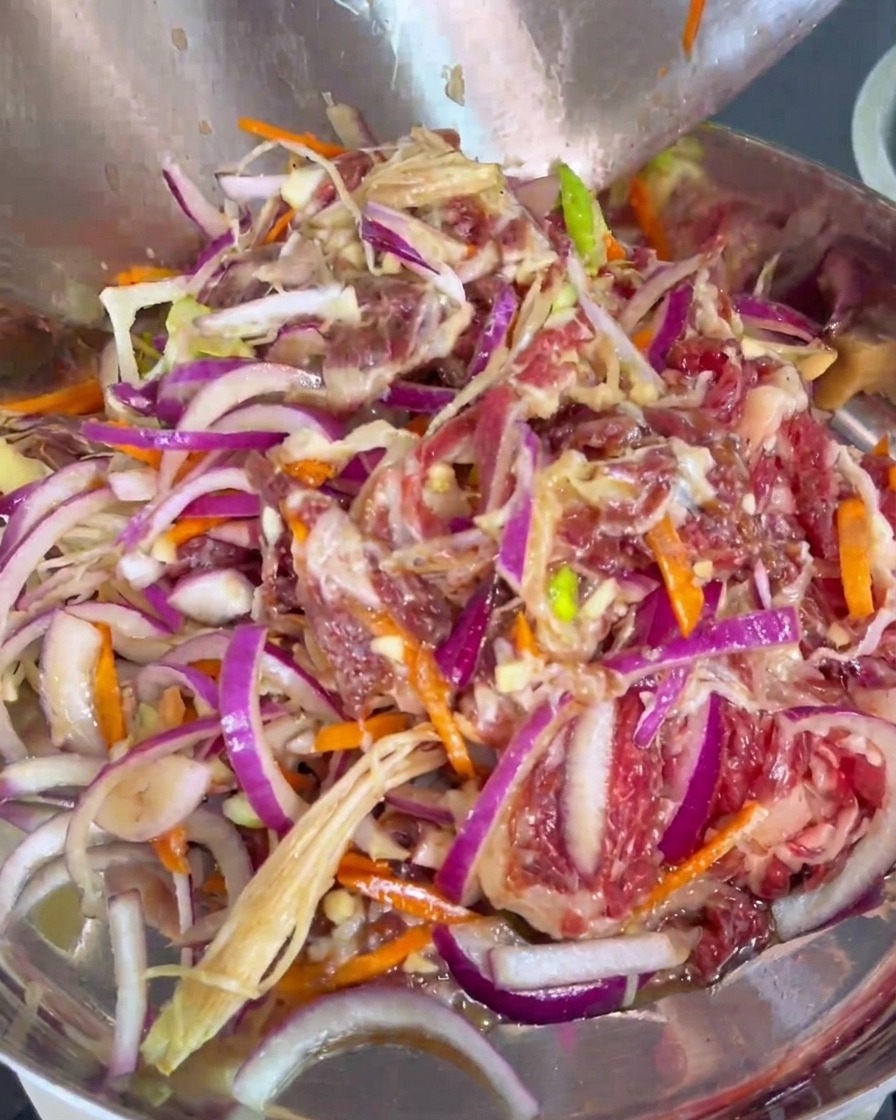 Tranches de bœuf mariné mélangées à des oignons rouges et des carottes transférées dans une poêle de cuisson en métal.