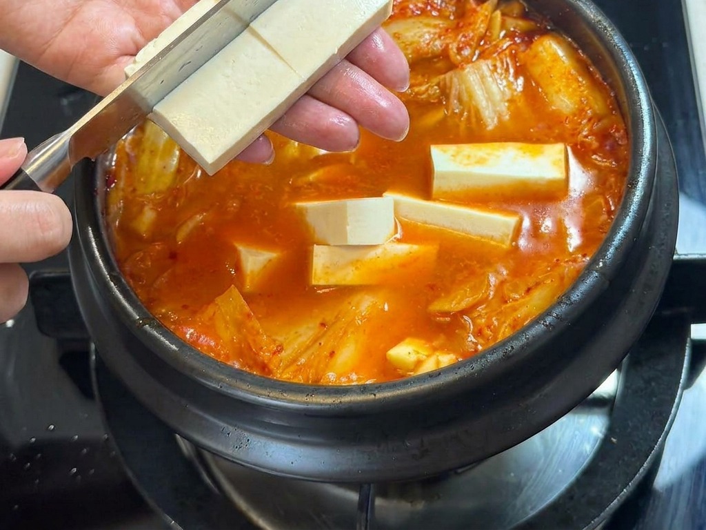 Une personne utilisant un couteau pour couper un bloc de tofu blanc directement au-dessus dun pot de ragoût de kimchi épicé.