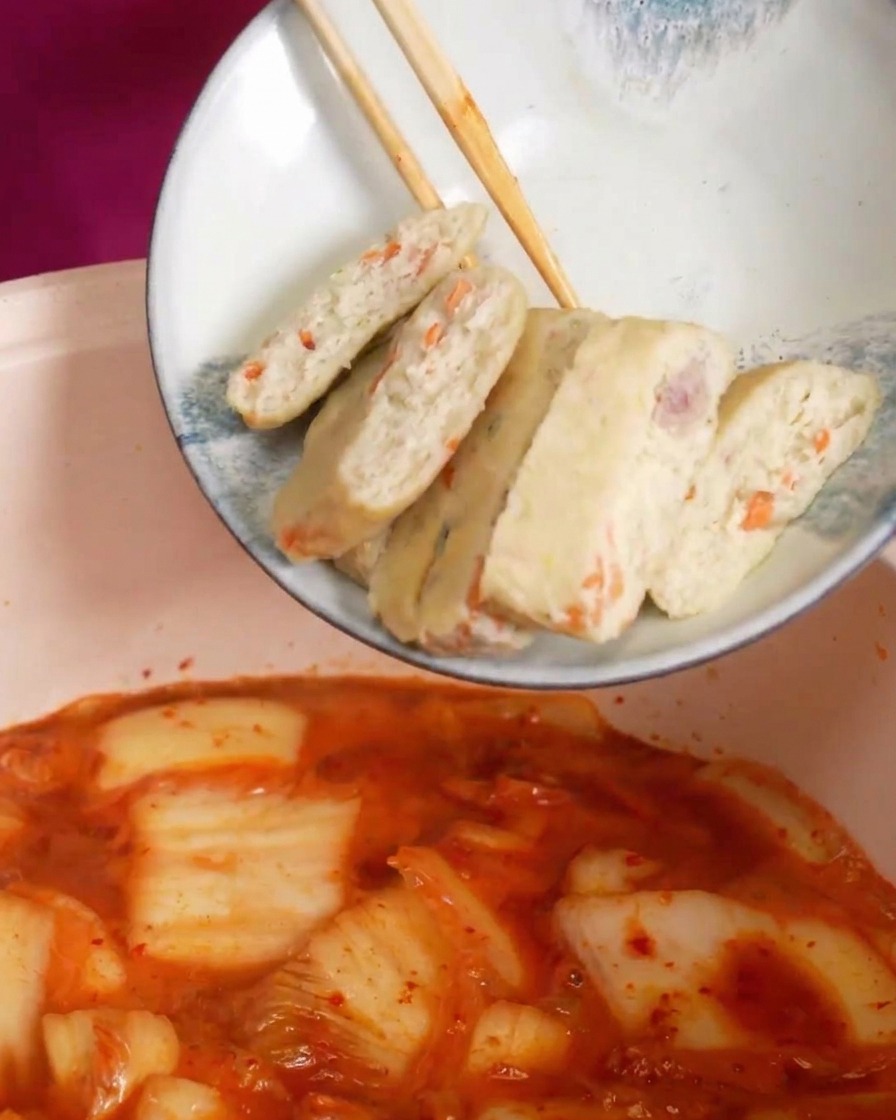 Tranches de gâteaux de poisson déposées dans une soupe de kimchi rouge et bouillonnante.