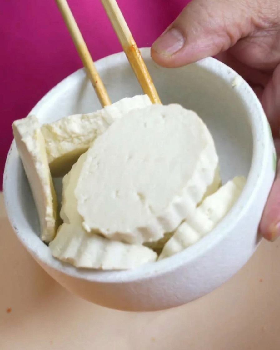 Bol blanc rempli de tranches de tofu blanc tenues au-dessus dune marmite de soupe rouge.