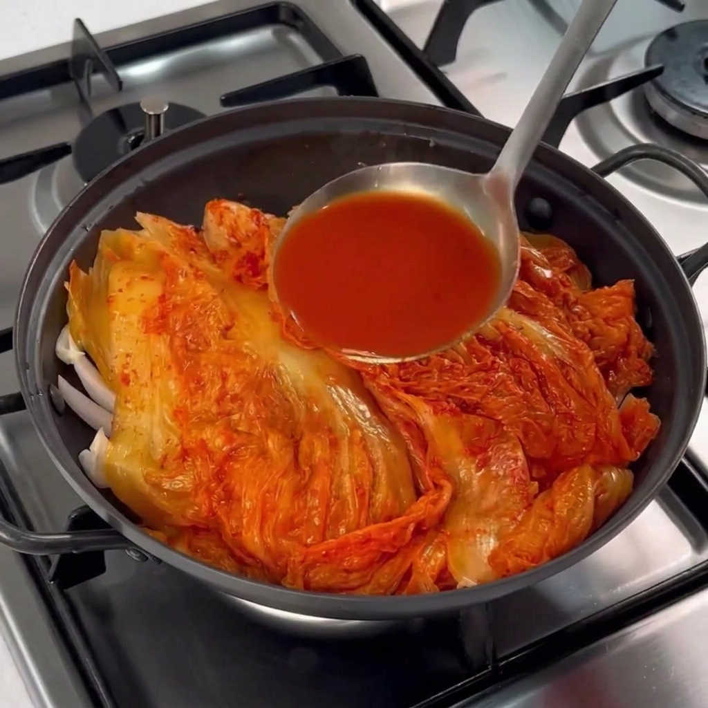 Une louche versant du jus de kimchi rouge vif sur le tas de kimchi et de porc dans une marmite noire.