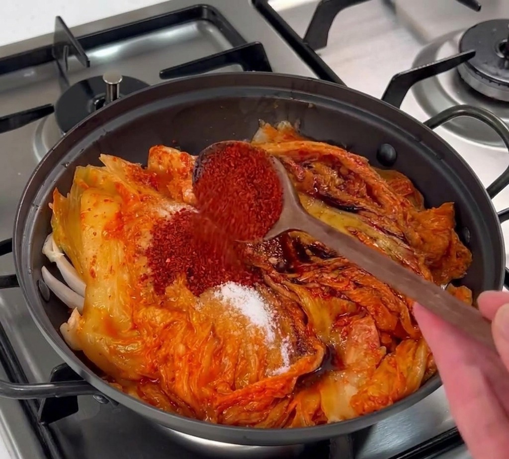 Une cuillère en bois versant des flocons de piment rouge sur la couche de kimchi.
