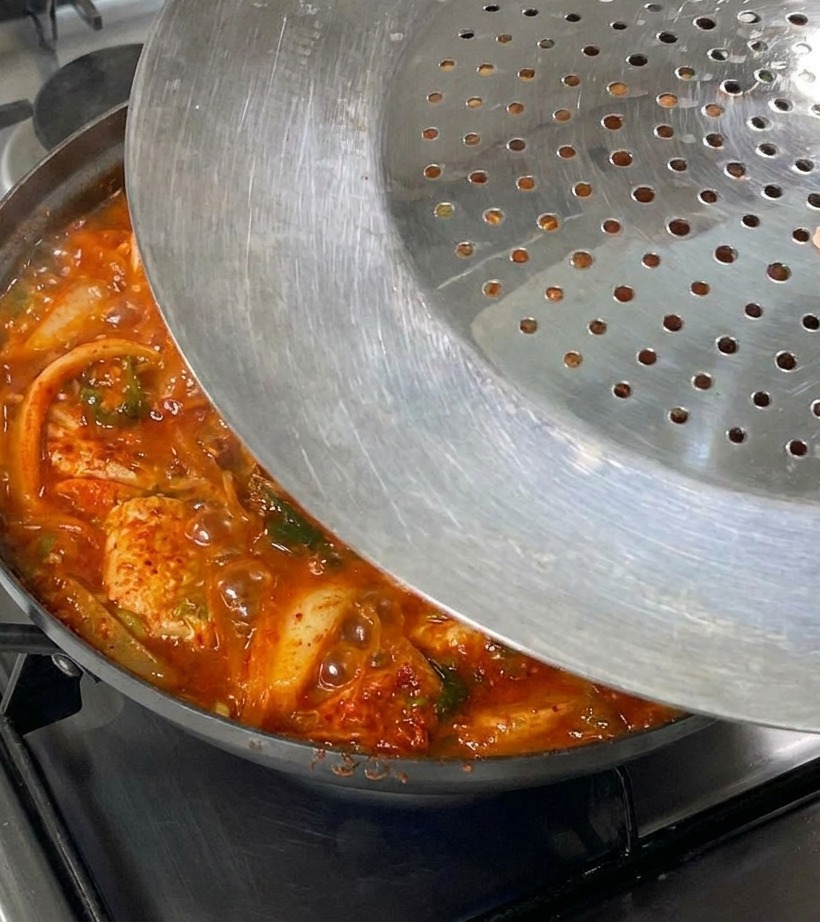 Une main posant un couvercle métallique sur une poêle bouillonnante de kimchi et de porc.