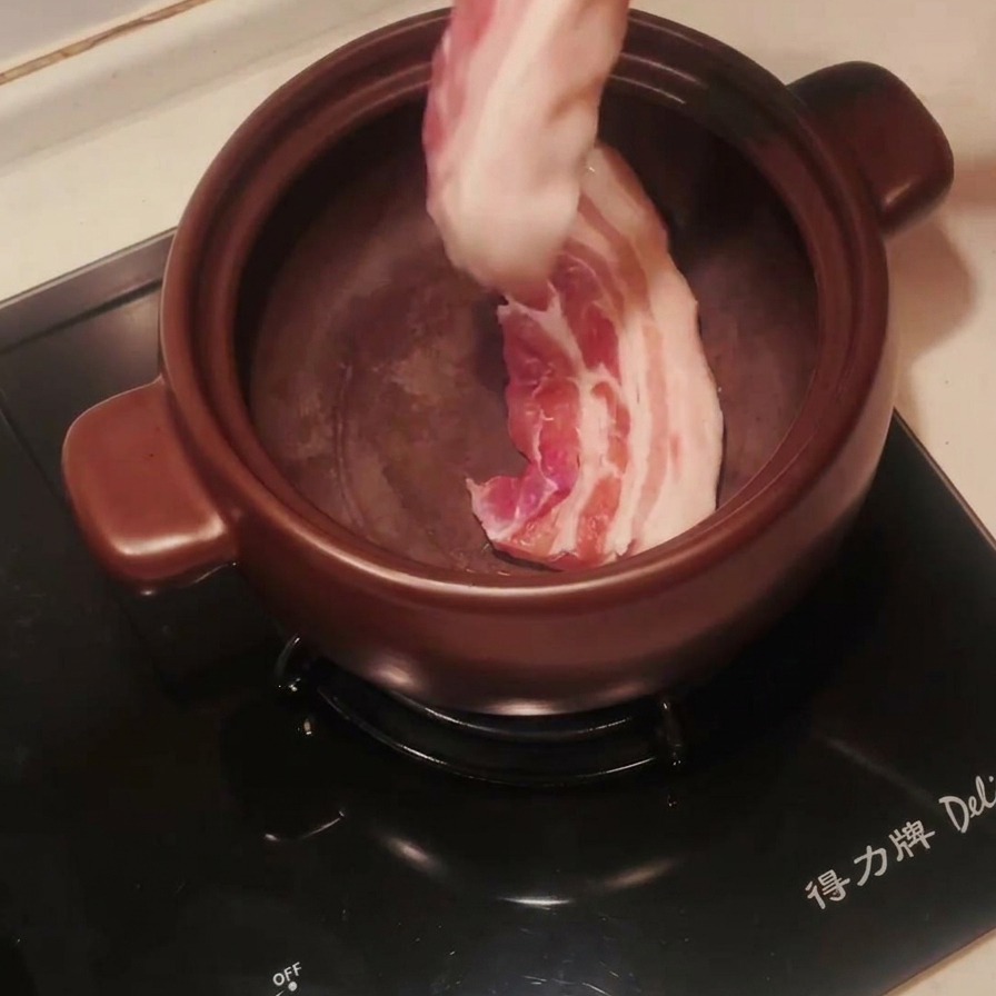 Tranches de poitrine de porc crues déposées avec une pince dans un pot en terre cuite préchauffé.