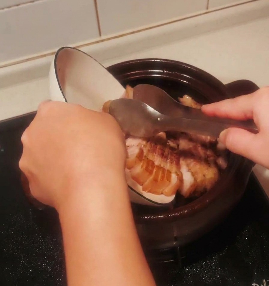 Morceaux de porc saisis versés dun bol blanc dans le pot en terre cuite sombre.