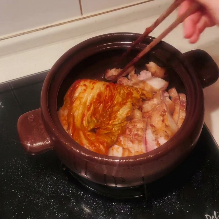 Une tête entière de kimchi assaisonné déposée sur les morceaux de porc dans le pot avec des baguettes.