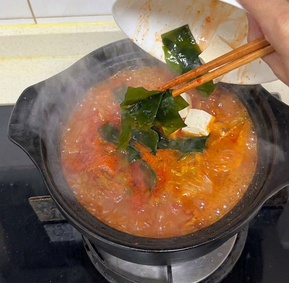 Une main versant des morceaux de varech vert foncé dans une marmite fumante de soupe rouge.