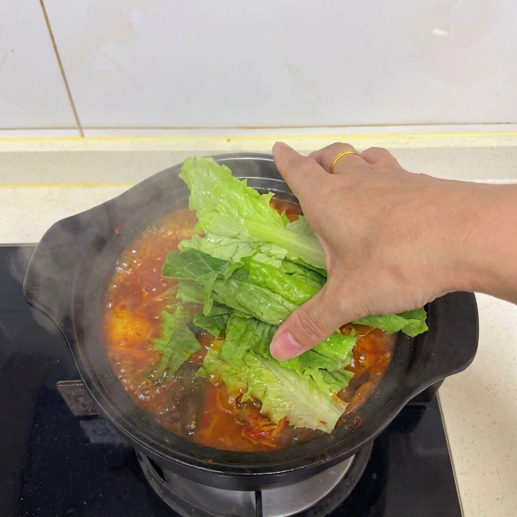 Une main plaçant des feuilles de laitue fraîches sur une soupe au kimchi fumante.
