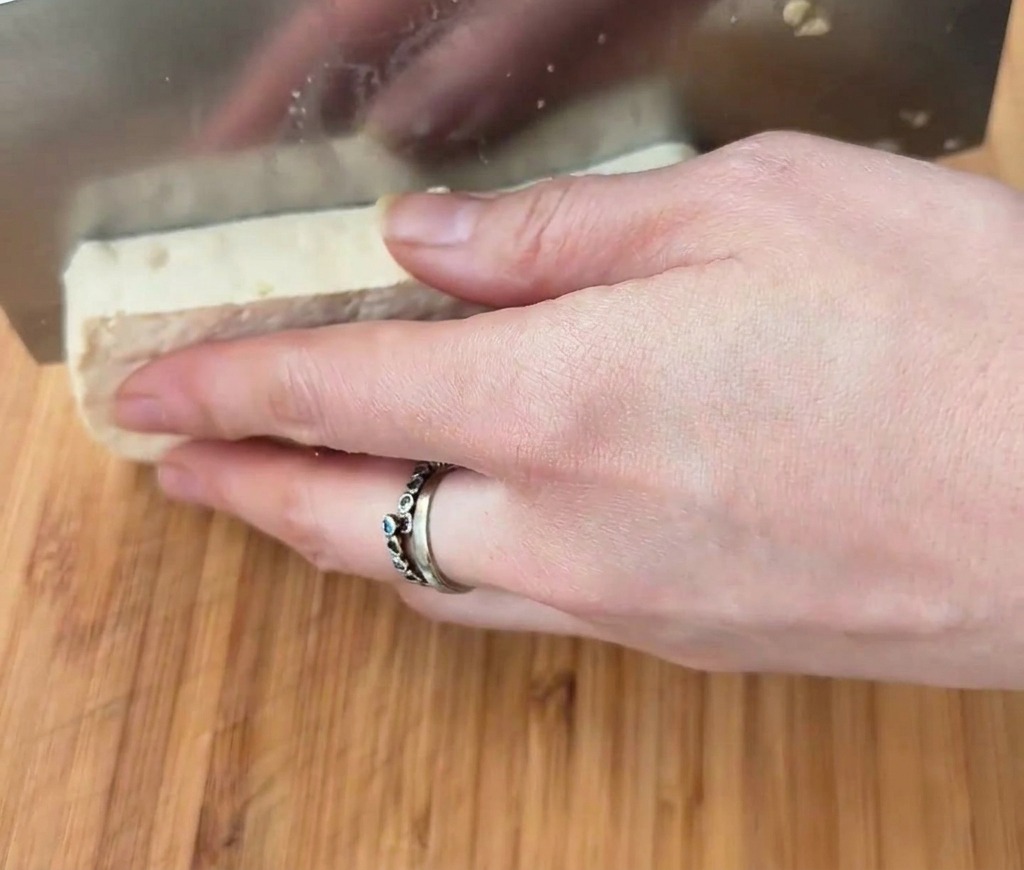 Bloc de tofu blanc coupé en cubes avec un large couperet sur une surface en bois.