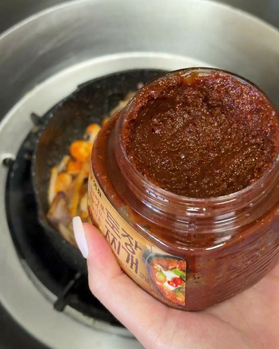 Une main tenant une cuillerée de pâte de soja coréenne foncée au-dessus dune poêle de légumes sautés.