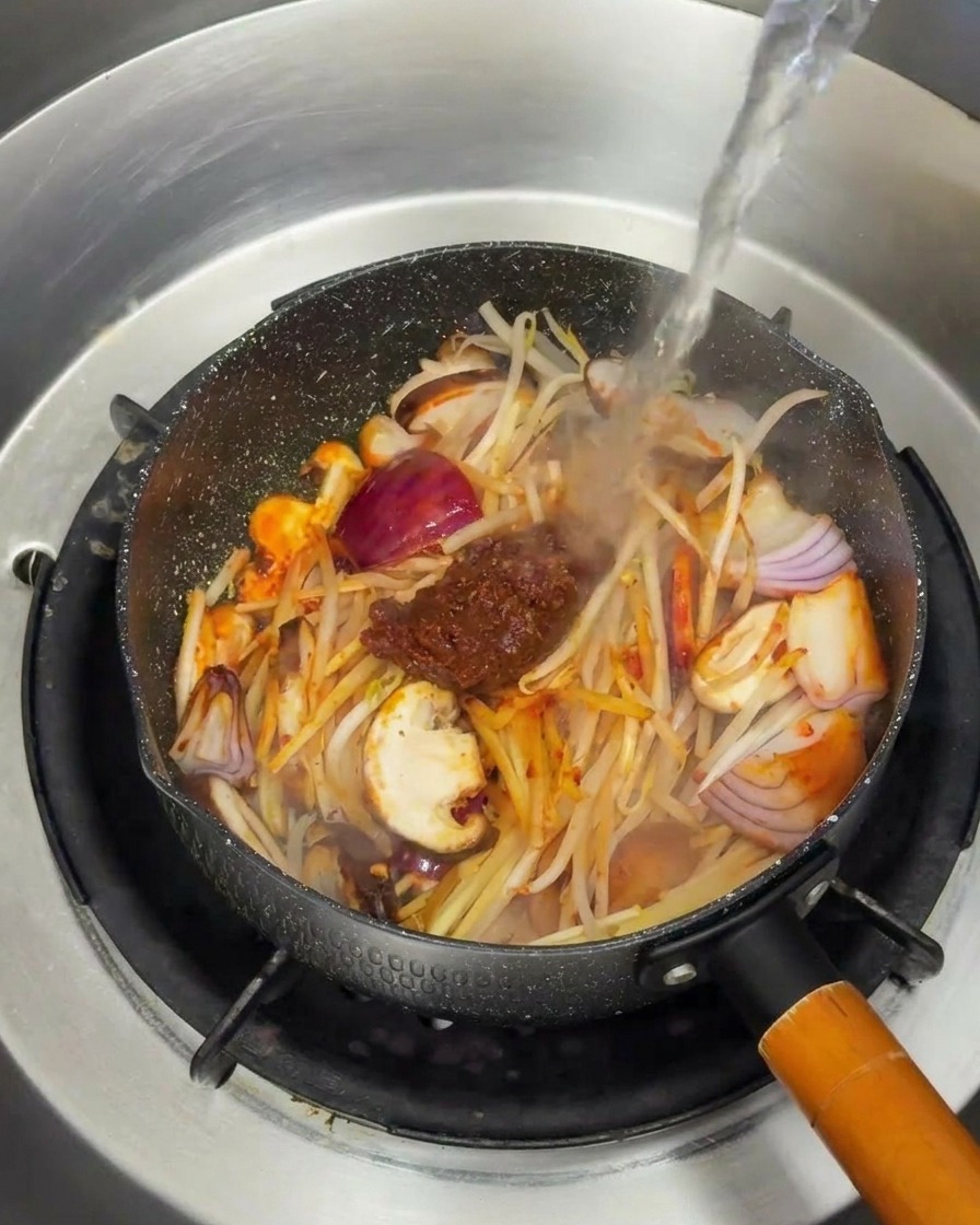 De leau bouillante versée sur les légumes sautés et la pâte de soja dans une poêle.