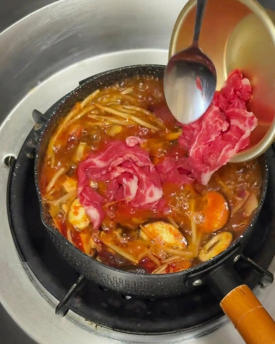 Fines tranches de bœuf cru ajoutées dans une marmite de bouillon rouge bouillonnant.