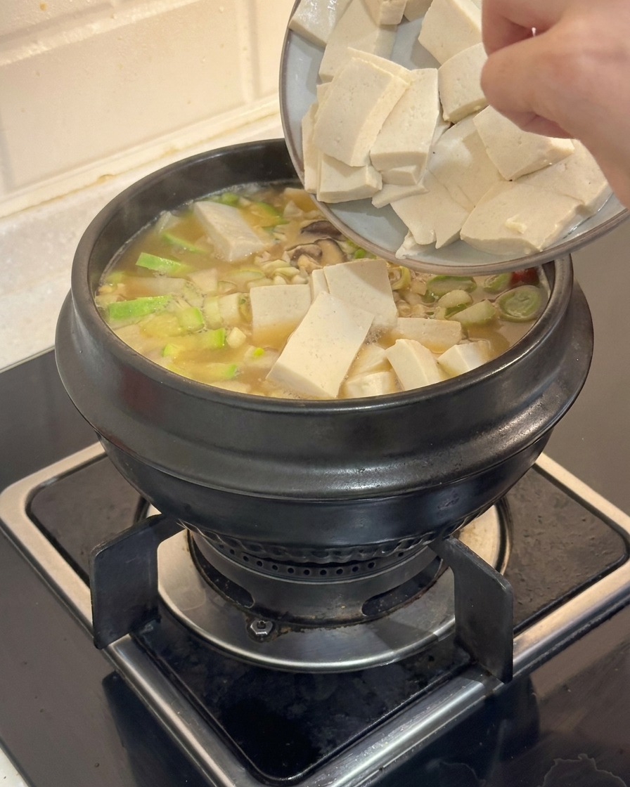 Une main faisant glisser des cubes de tofu blanc dune assiette dans un ragoût coréen fumant