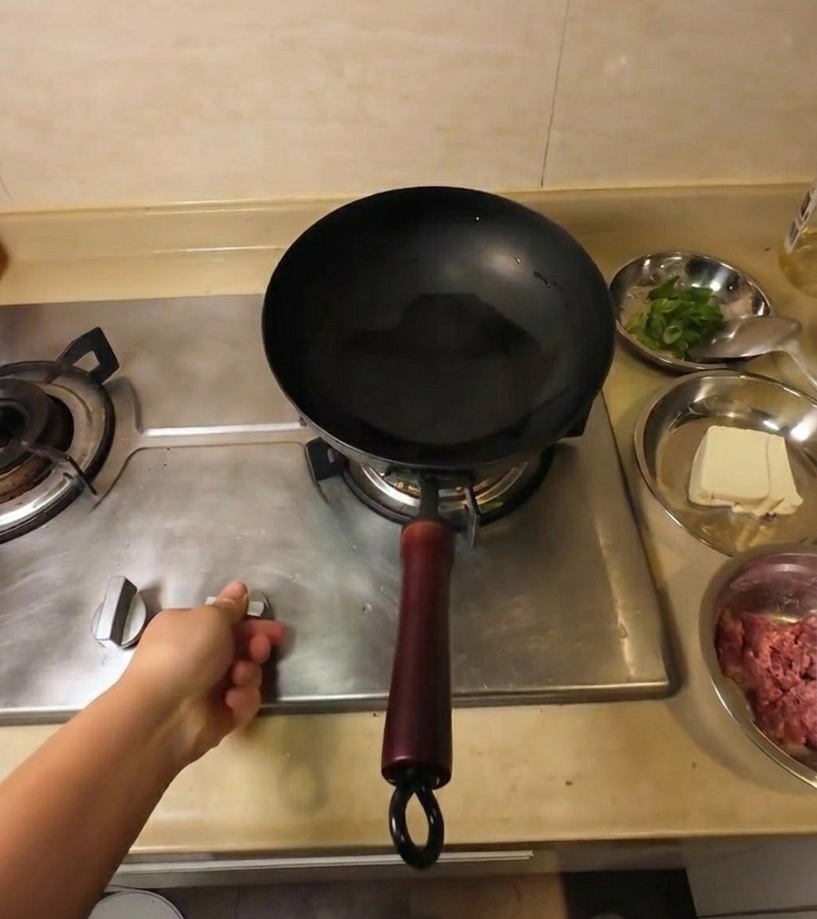 Une main réglant le bouton dune cuisinière à gaz sous un wok noir contenant de lhuile de cuisson limpide.