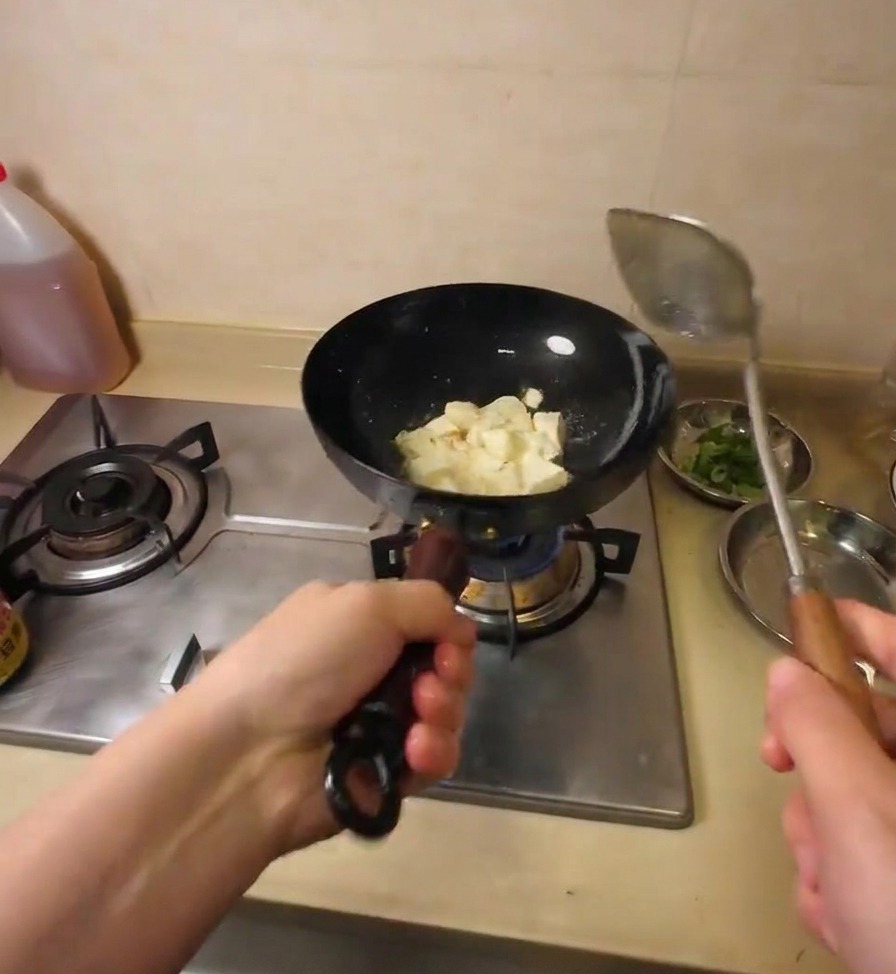 Une spatule métallique cassant un gros bloc de tofu en cubes irréguliers pendant quil grésille dans lhuile chaude.