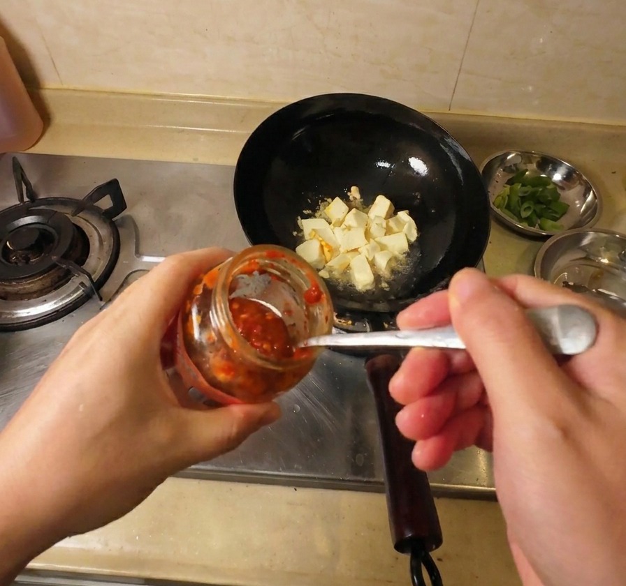 Gros plan dune main utilisant une cuillère métallique pour ajouter de la sauce chili rouge dun bocal en verre dans un wok rempli de morceaux de tofu frit.