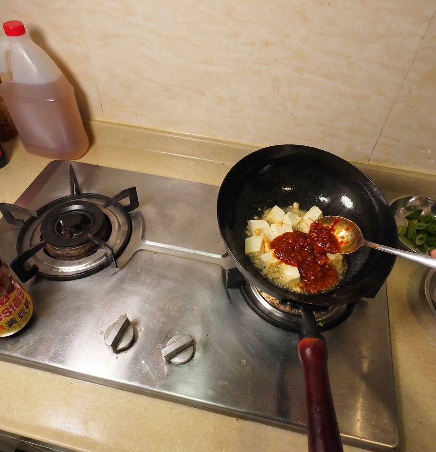 Pâte de soja brun foncé sur une cuillère métallique ajoutée dans le wok à côté du tofu frit et de la sauce chili rouge.