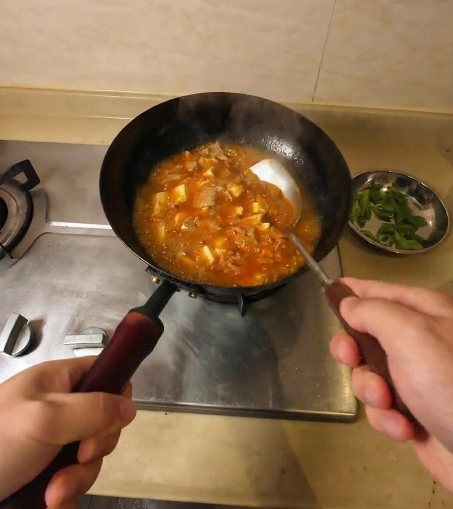 Une personne remuant une soupe épaisse orange-rouge bouillonnante contenant du tofu et du bœuf dans un wok noir.
