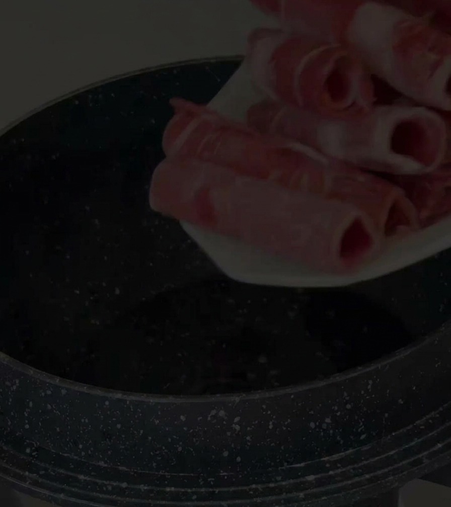 Une main déposant de fines tranches de poitrine de bœuf crue dans une marmite antiadhésive noire.