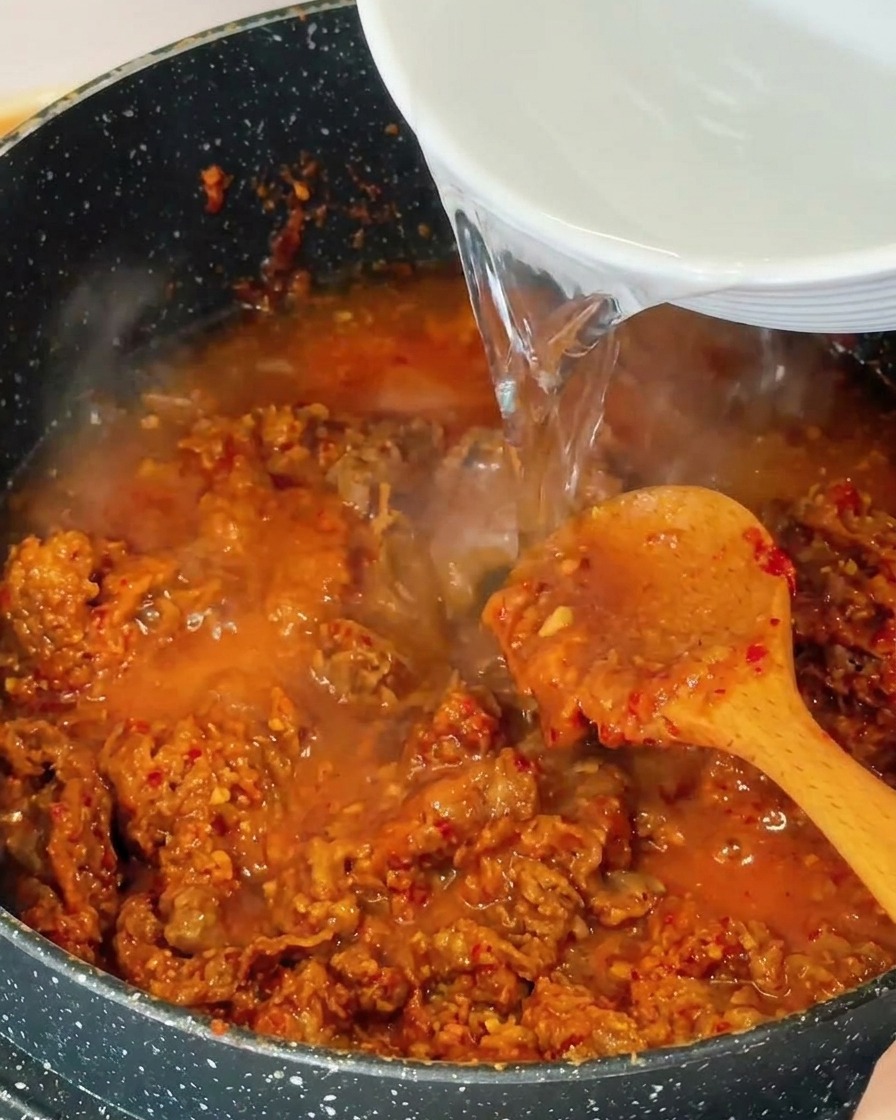 Eau claire versée depuis un bol blanc dans une casserole contenant le bœuf sauté et un bouillon bouillonnant.