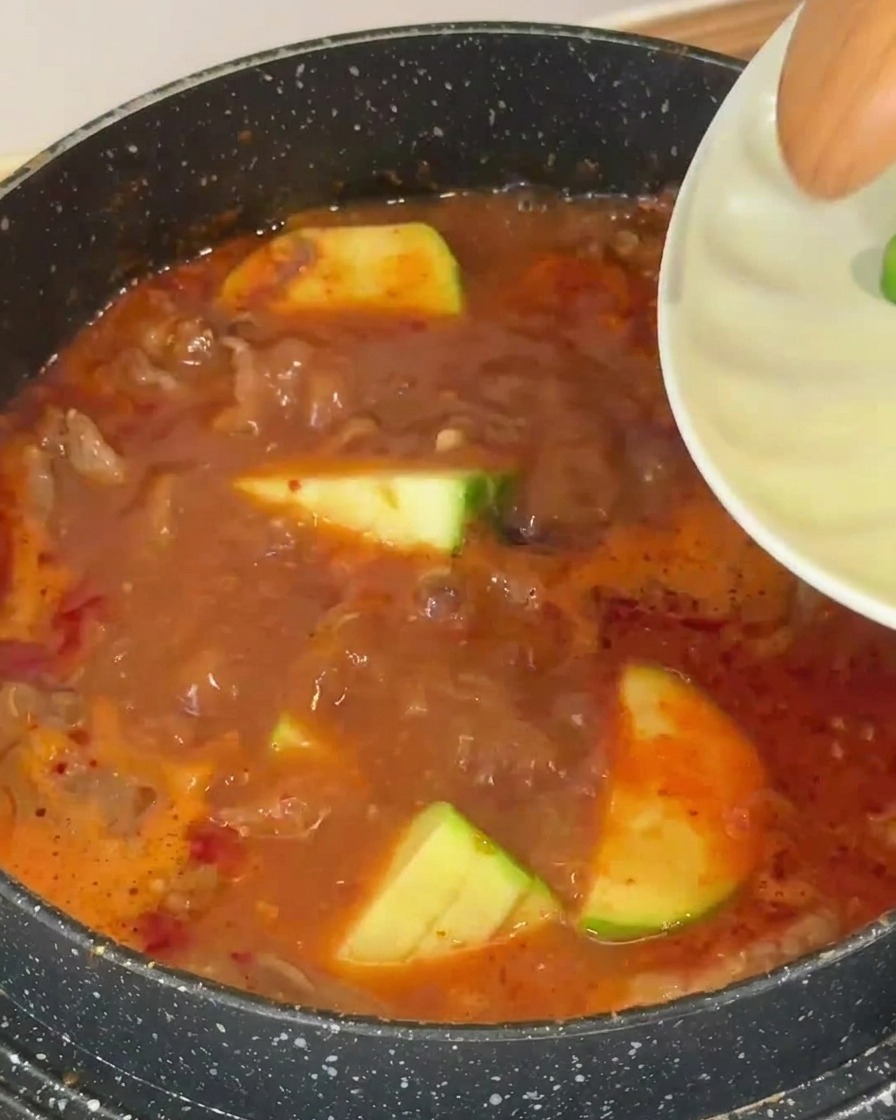 Plusieurs tranches de courgette à la peau verte ajoutées dans un bouillon de soupe rouge-brun bouillonnant.