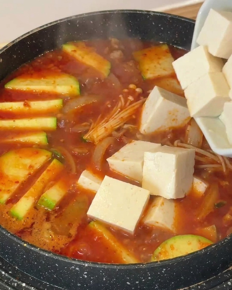 Cubes de tofu blanc ajoutés dans une marmite contenant la soupe, les courgettes et les champignons enoki.