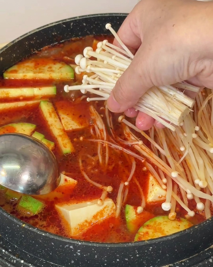 Une main plaçant un bouquet de longs et fins champignons enoki dans une soupe bouillante avec tofu et courgettes.