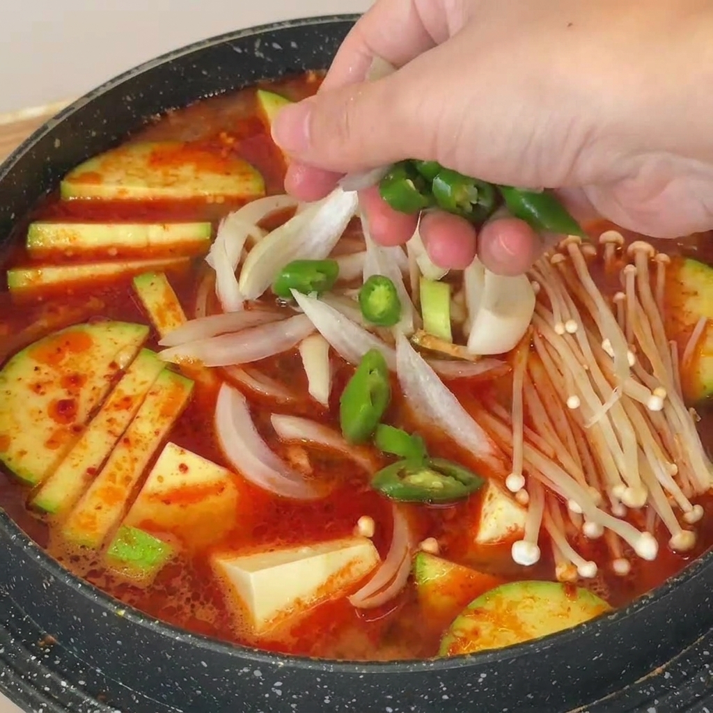 Main saupoudrant des tranches de piment vert sur une soupe de pâte de soja coréenne garnie de champignons et tofu.