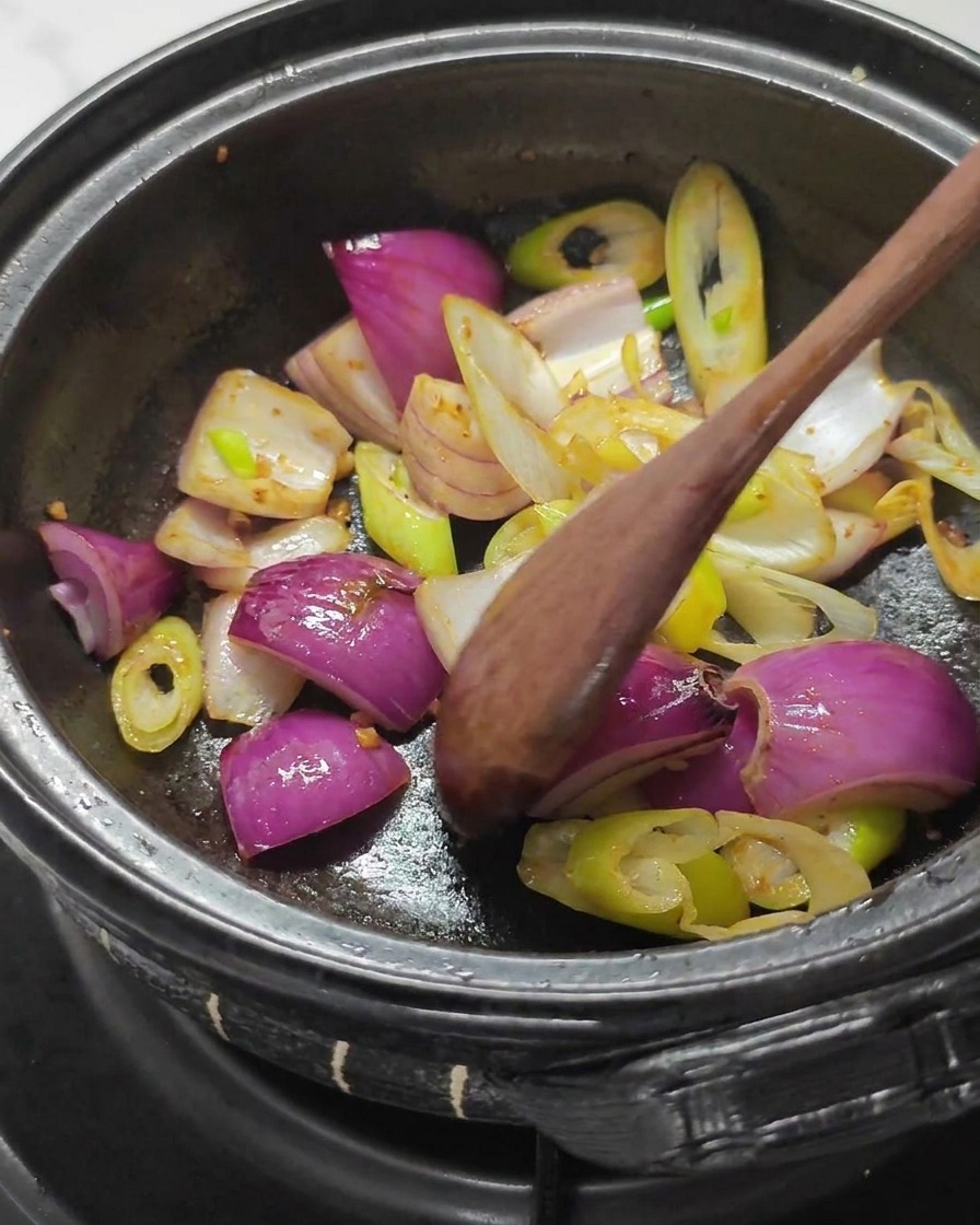 Cuillère en bois mélangeant de gros morceaux doignon rouge et de blanc doignon vert dans une marmite en pierre sombre.
