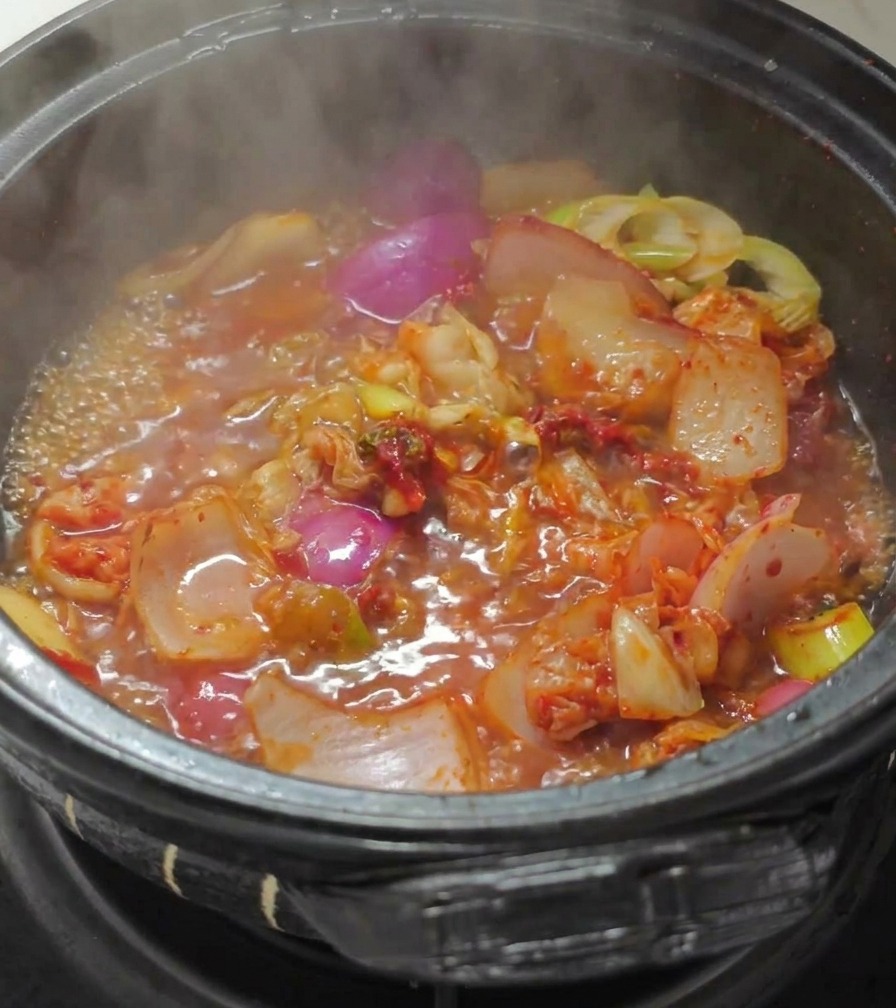 Eau bouillante versée dans une marmite en pierre noire contenant du kimchi sauté et des oignons dans un bouillon rouge vibrant.