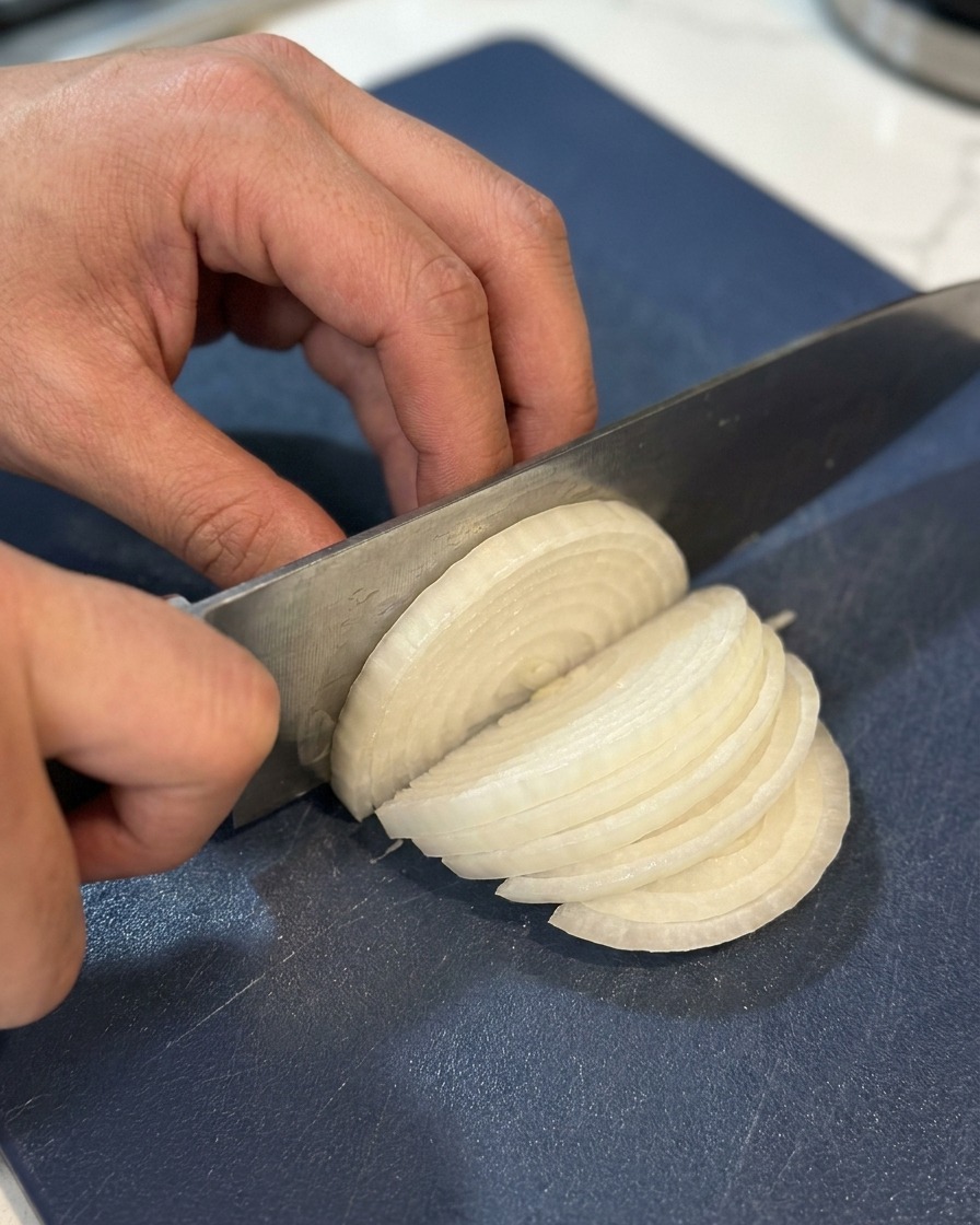 Gros plan de mains utilisant un couteau de chef pour émincer un demi-oignon blanc en fines lamelles sur une planche à découper bleu marine.