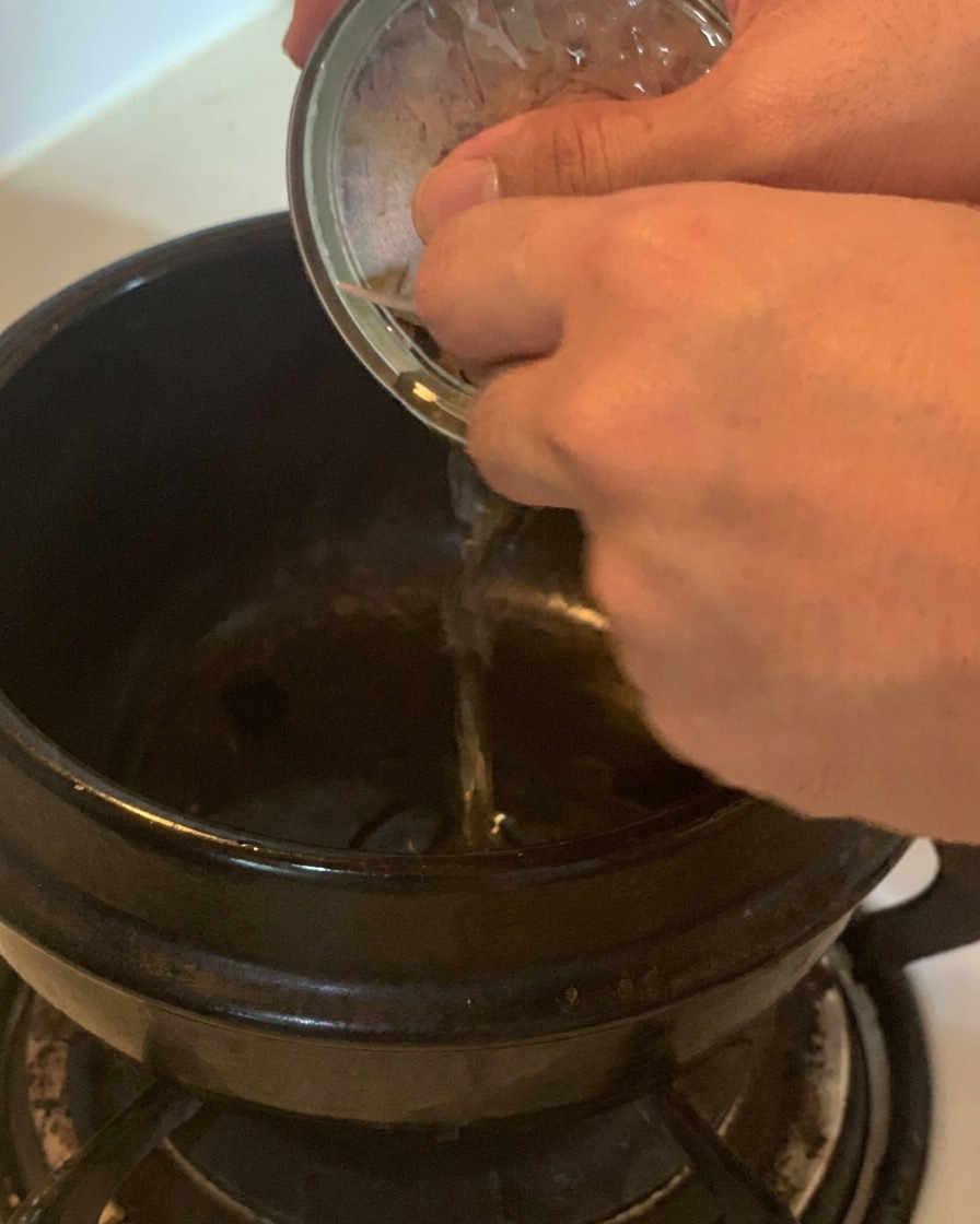 Une main versant lhuile dune boîte de thon argentée dans un faitout en céramique noire sur une cuisinière.