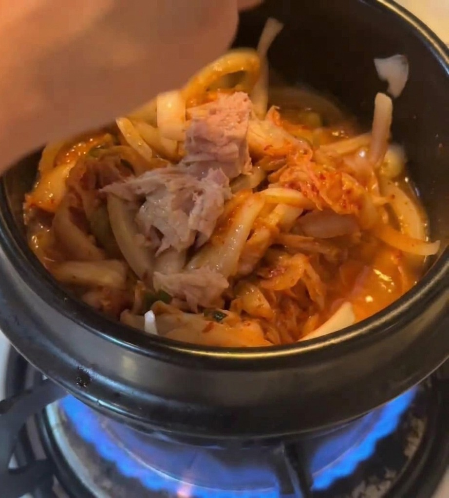 Morceaux de thon en conserve ajoutés dans un pot contenant le kimchi et les oignons sur une flamme de gaz bleue.