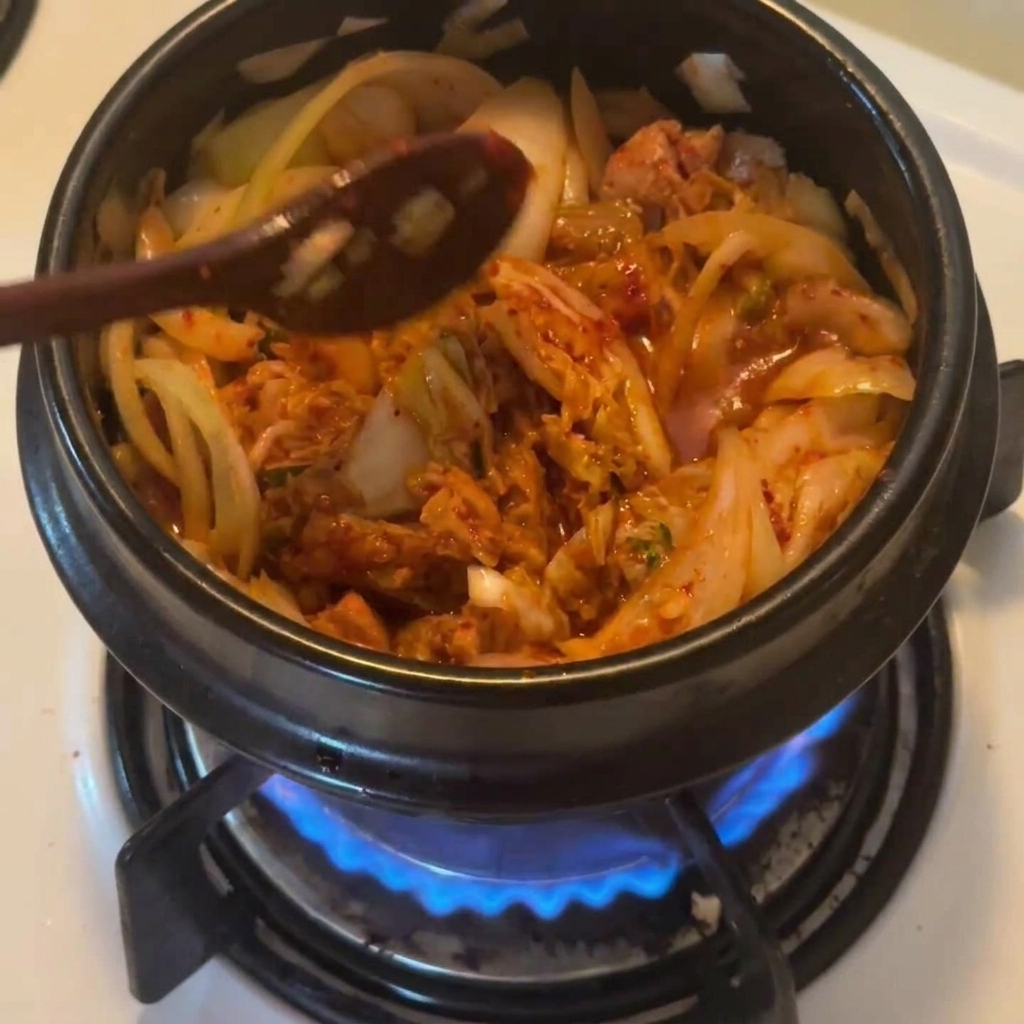 Une cuillère en bois saupoudrant du piment coréen rouge sur le mélange thon-kimchi dans un pot noir.