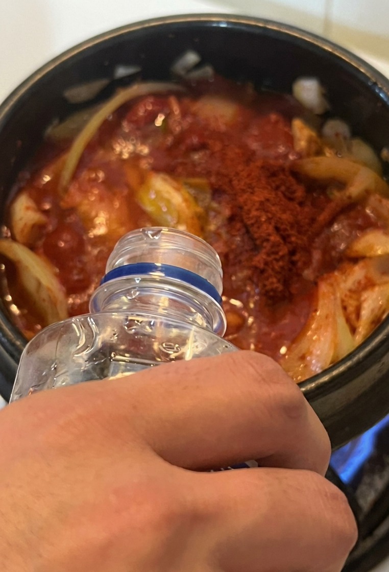 Eau versée dune bouteille transparente dans un pot de ragoût rouge sur une cuisinière à gaz.