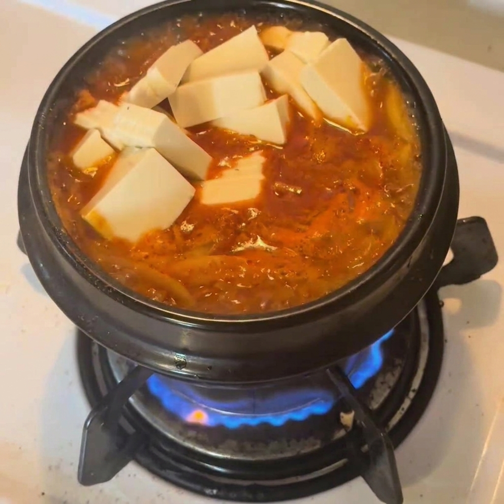 Cubes de tofu blanc frais posés sur un ragoût de thon au kimchi rouge vif dans un pot en céramique noire.